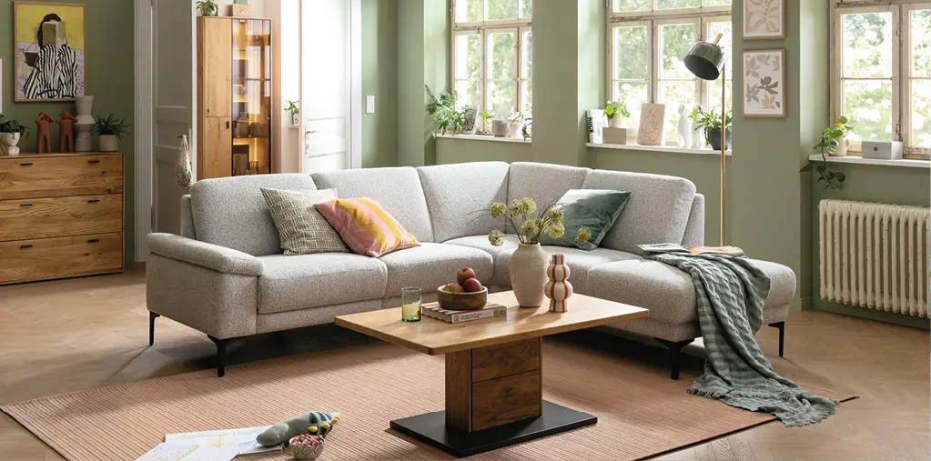 Helles Wohnzimmer mit großem Ecksofa in Stoffoptik, Holzcouchtisch und grüner Wandfarbe. Natürliche Einrichtung mit Wohnaccessoires, Pflanzen und viel Tageslicht für ein modernes und gemütliches Wohnambiente.